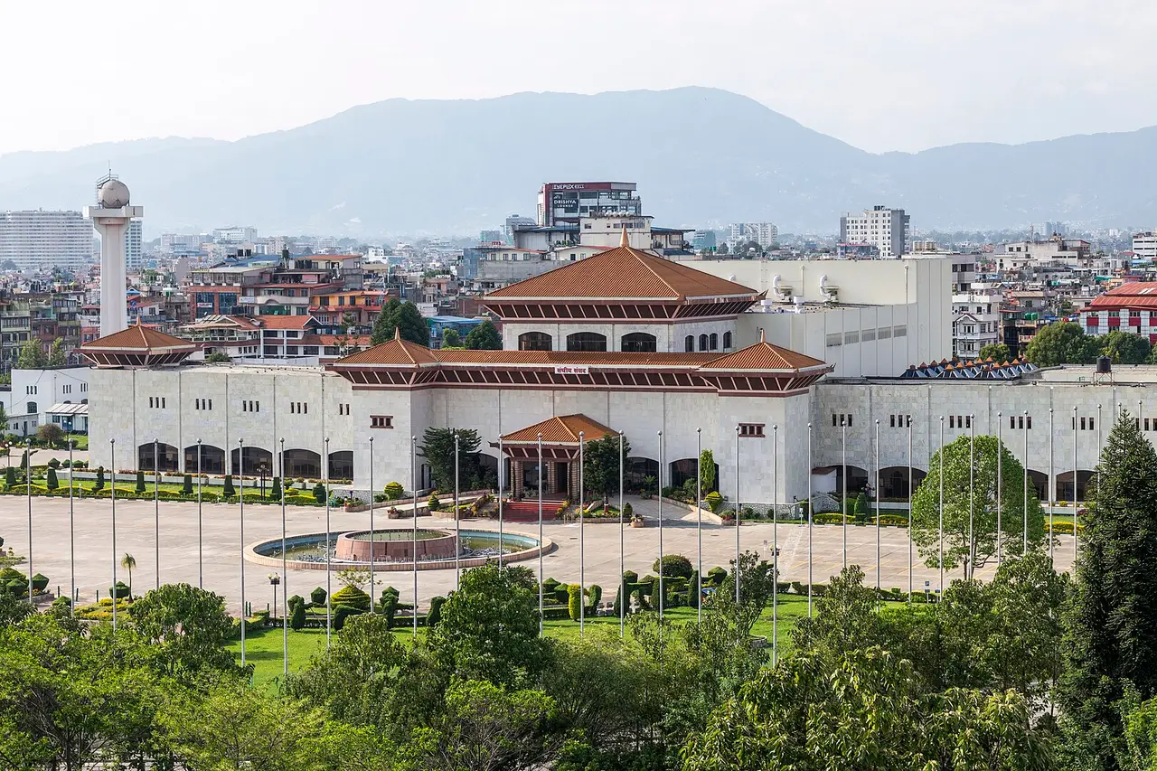 A large white government building with tiered red roofs, gardens in front and hills behind